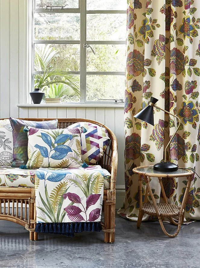 A cozy sitting area with a rattan sofa decorated with colorful botanical patterned pillows and throw. To the right, a wooden side table with a black desk lamp and a wicker chair. In the background, a window with a potted plant on the sill and floral 