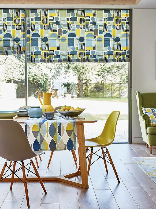 A dining area with a wooden table set with bowls, a pitcher, and cups, surrounded by four chairs in different colors; a window with patterned curtains reveals a backyard with trees and grass.