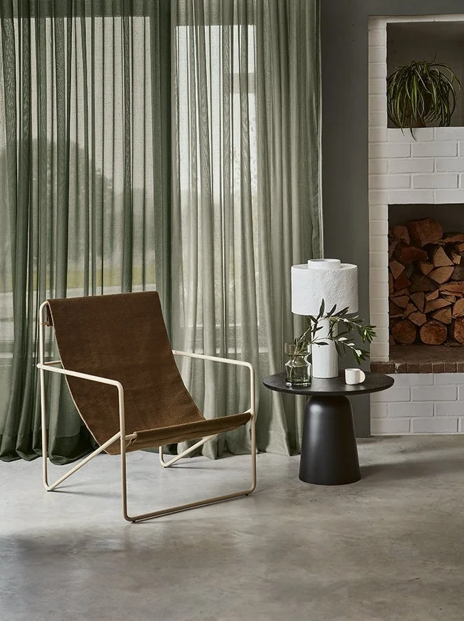 A living room corner featuring a brown recliner chair with white metal armrest and frame, a black round side table with a white vase holding green foliage, a white table lamp, and a fireplace with stacked firewood and a potted plant on the mantle. Gr
