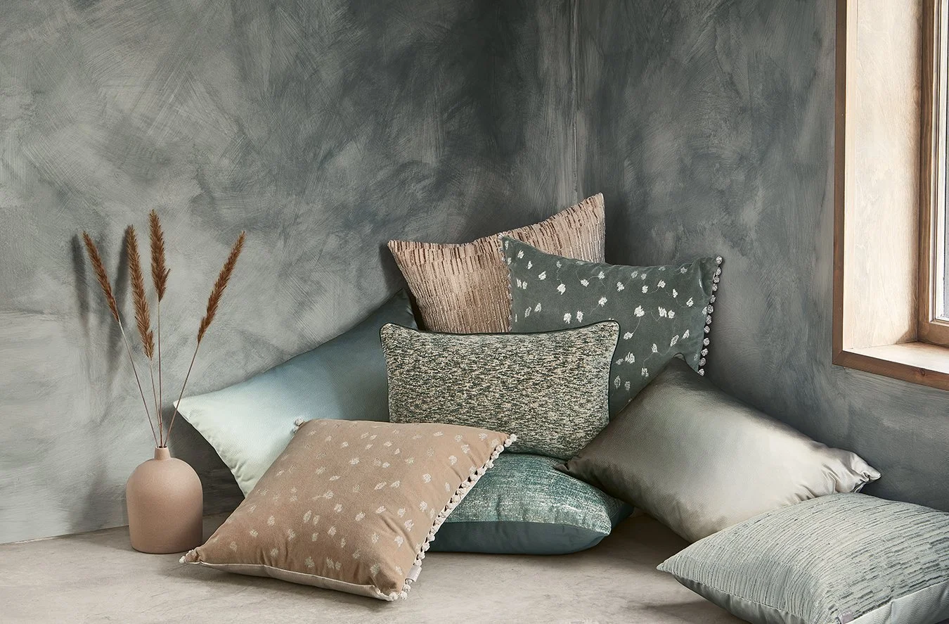 A cozy corner with a variety of decorative pillows in muted colors and patterns, beside a beige vase with dried grasses, by a window with natural light.