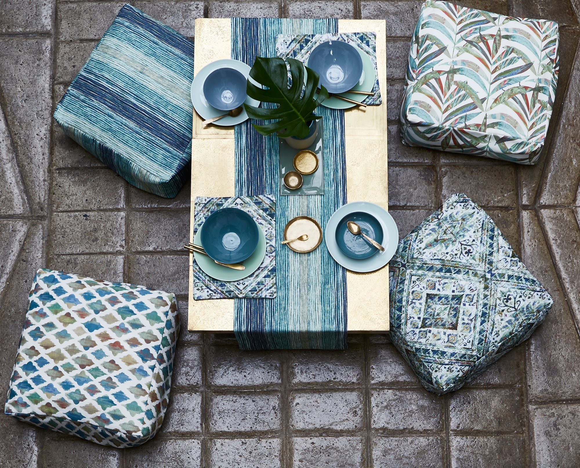 A top-down view of a table set for a meal, surrounded by four cushioned seats with colorful patterns, on a stone patio. The table has blue and gold dishes, bowls, and utensils, with a centerpiece of a green leaf in a white vase.