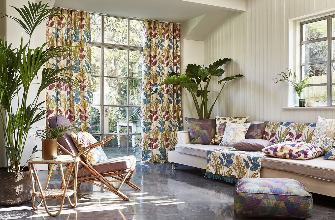 Living room with large windows, floral curtains, and a white sofa with patterned cushions, surrounded by green houseplants, including large leaves and potted plants.
