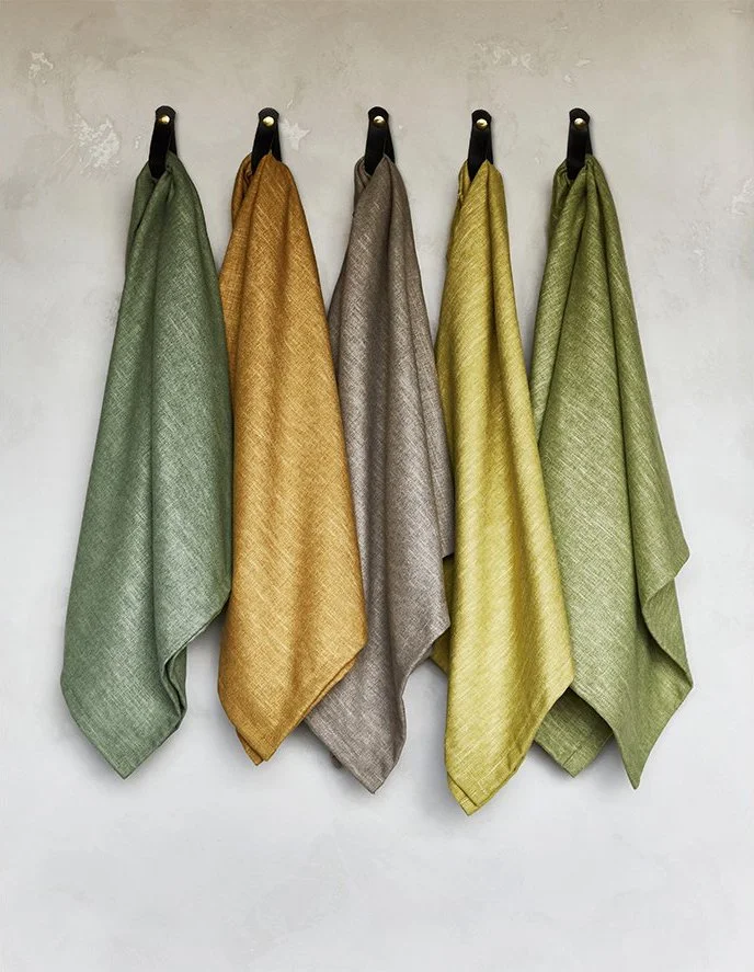 Five hanging cloths in pastel colors, including green, orange, gray, yellow, and light green, on black hooks against a plain wall.