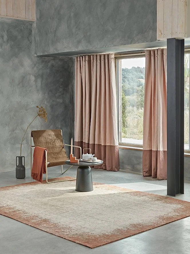 A cozy living room corner featuring a brown armchair with a red throw blanket, a black side table with a teapot and cups, a gray rug, pink curtains, a large window with a view of green trees, and a gray textured wall.