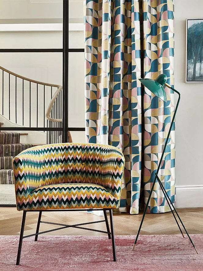 A colorful patterned armchair, a tall teal floor lamp, patterned curtains, and a pink rug in a modern interior