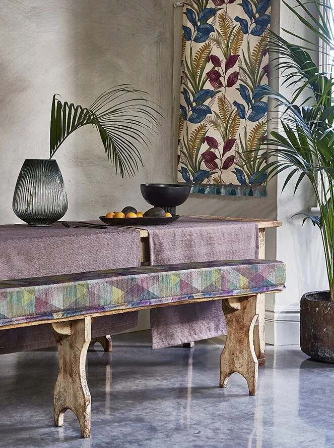 Interior of a room with a wooden bench with a colorful, geometric-patterned cushion, a lavender covering, and a rustic table decorated with a black vase holding palm leaves, a black bowl with lemons, and a fabric wall hanging with botanical patterns 