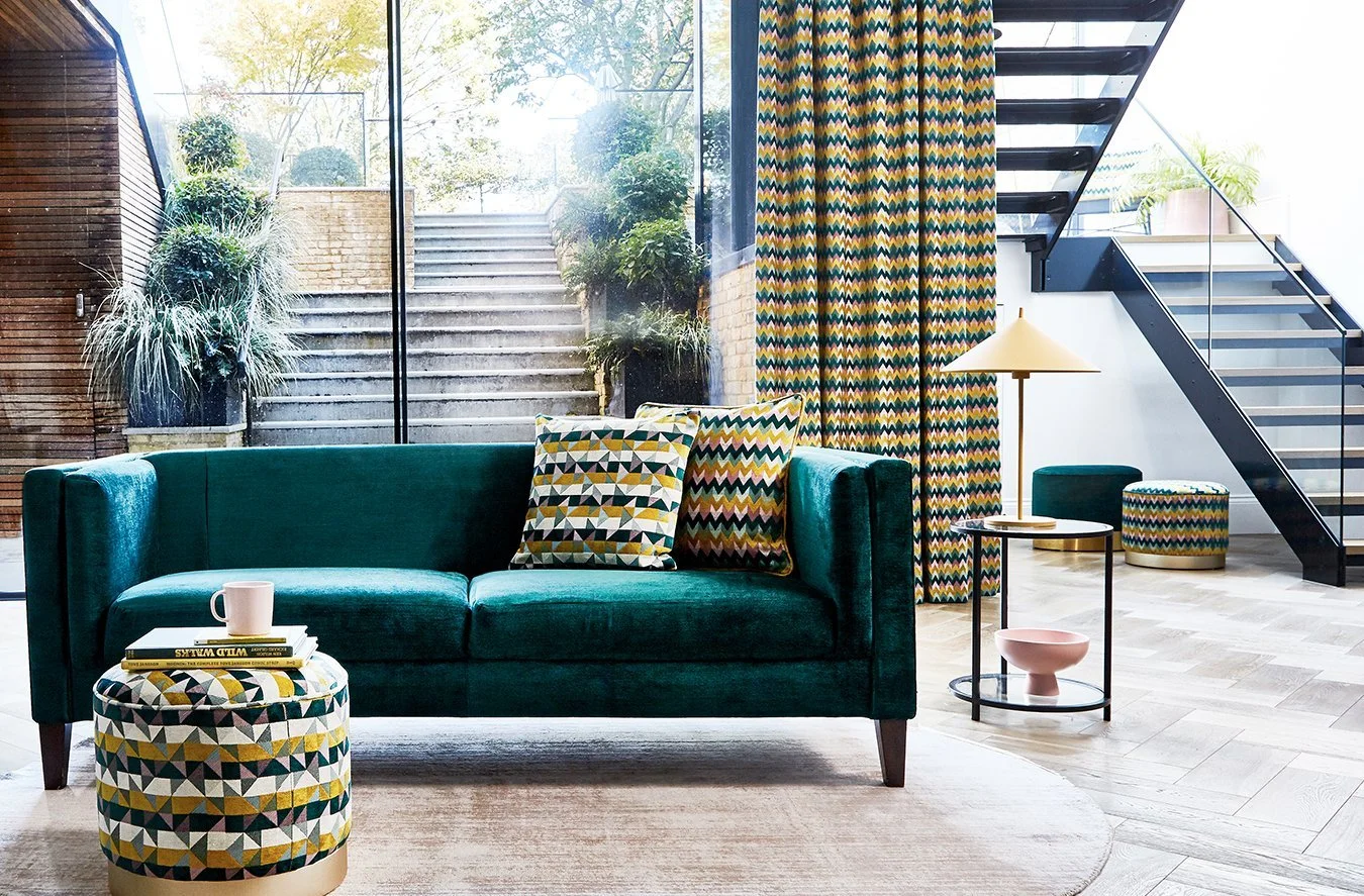 Living room with teal sofa, colorful patterned cushions, round patterned ottoman, side table with lamp, large window with brick and indoor plants outside, colorful patterned curtains, staircase with black railing.