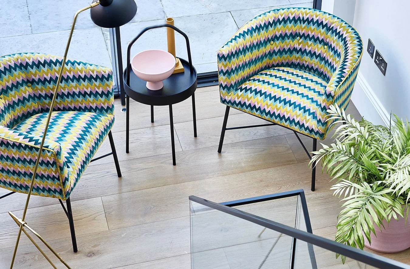 Colorful patterned armchairs with yellow, blue, and pink zigzag designs, a small black side table with a pink ceramic bowl, a black and yellow lamp, a plant in a pink pot, and a glass railing near a large window.