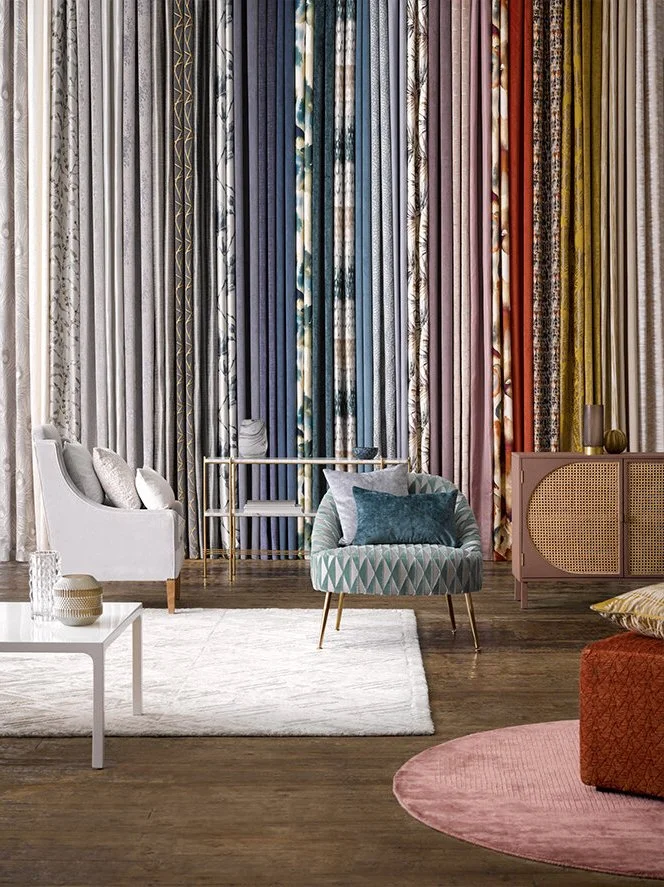 A living room with colorful curtains, a white sofa with pillows, a patterned blue armchair with pillows, a white coffee table with a vase, a pink sideboard with decorative items, and a pink rug on a wooden floor.