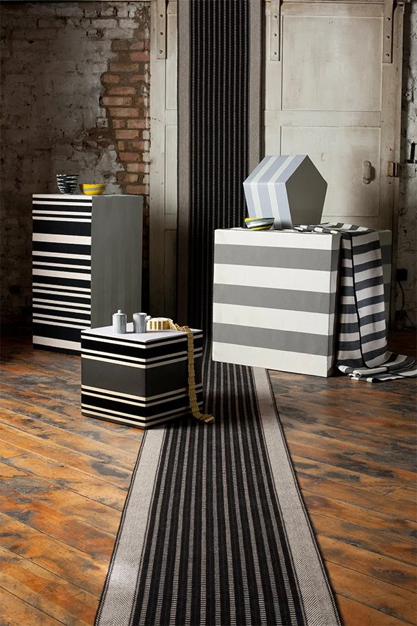 Interior featuring three striped storage units with a black and white striped rug on a wooden floor. Decorative bowls and small jars are on top of the units, and a striped cloth drapes over one. The background includes an exposed brick wall and verti