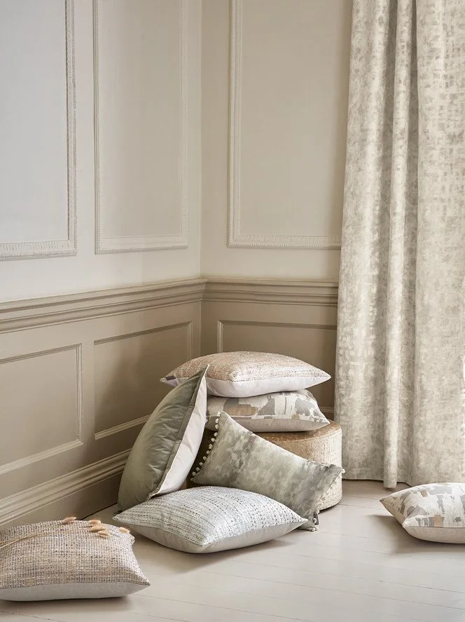 A corner of a room with beige wainscoting walls, a small cushioned stool, and a cluster of decorative pillows on the floor and stool near beige curtains.