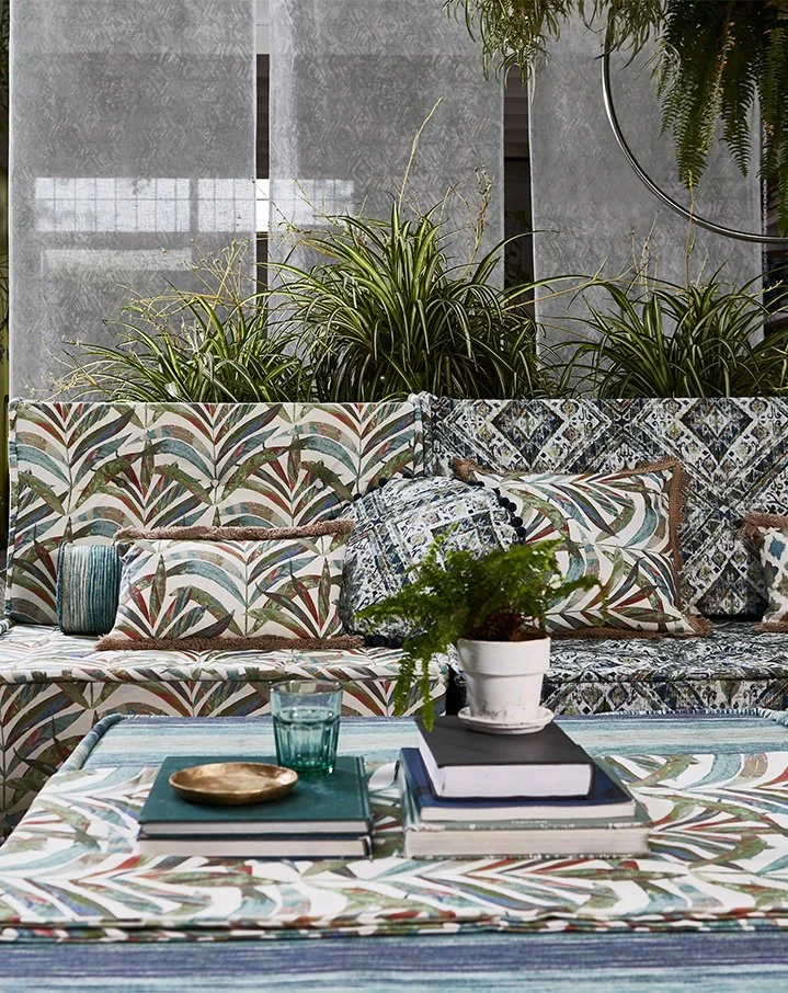 Interior with patterned sofa, cushions, a table with books, a potted fern, and green plants in the background.