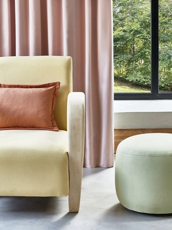 A living room corner with a yellow upholstered armchair with a rust-colored pillow, soft pink curtains, and a window showing green trees outside. Next to the armchair is a light green round ottoman.