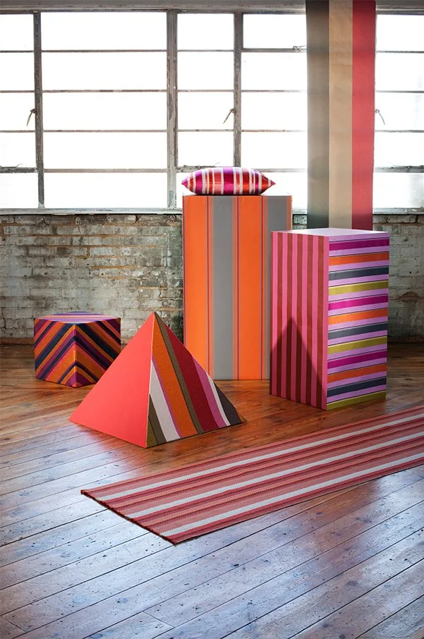 Colorful striped pedestals and a pillow arranged in a room with wooden flooring and large industrial-style windows.