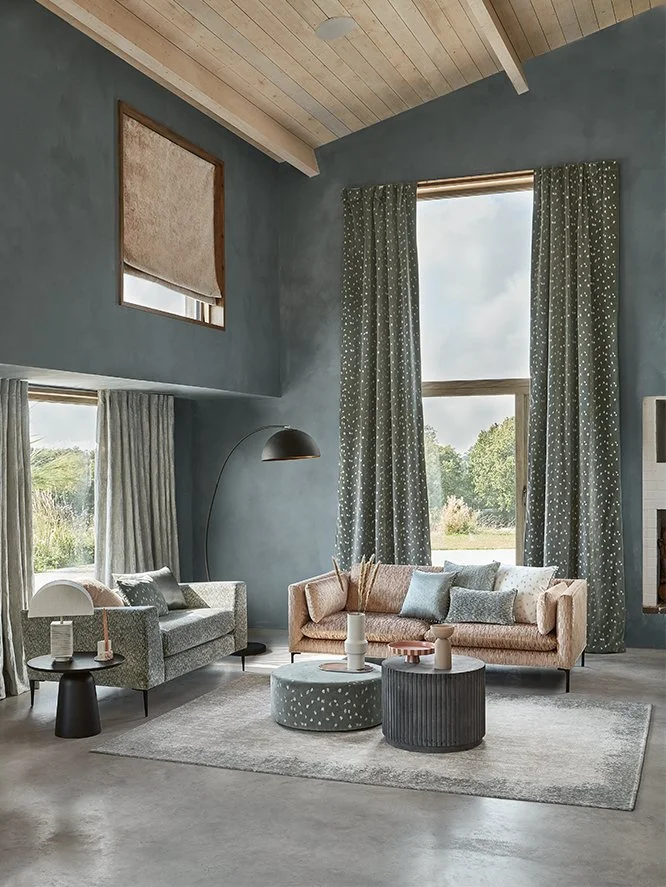 Living room with light blue walls, high vaulted ceiling with wood paneling, large windows with patterned curtains, two sofas in pastel tones, round coffee tables, and decorative pillows.