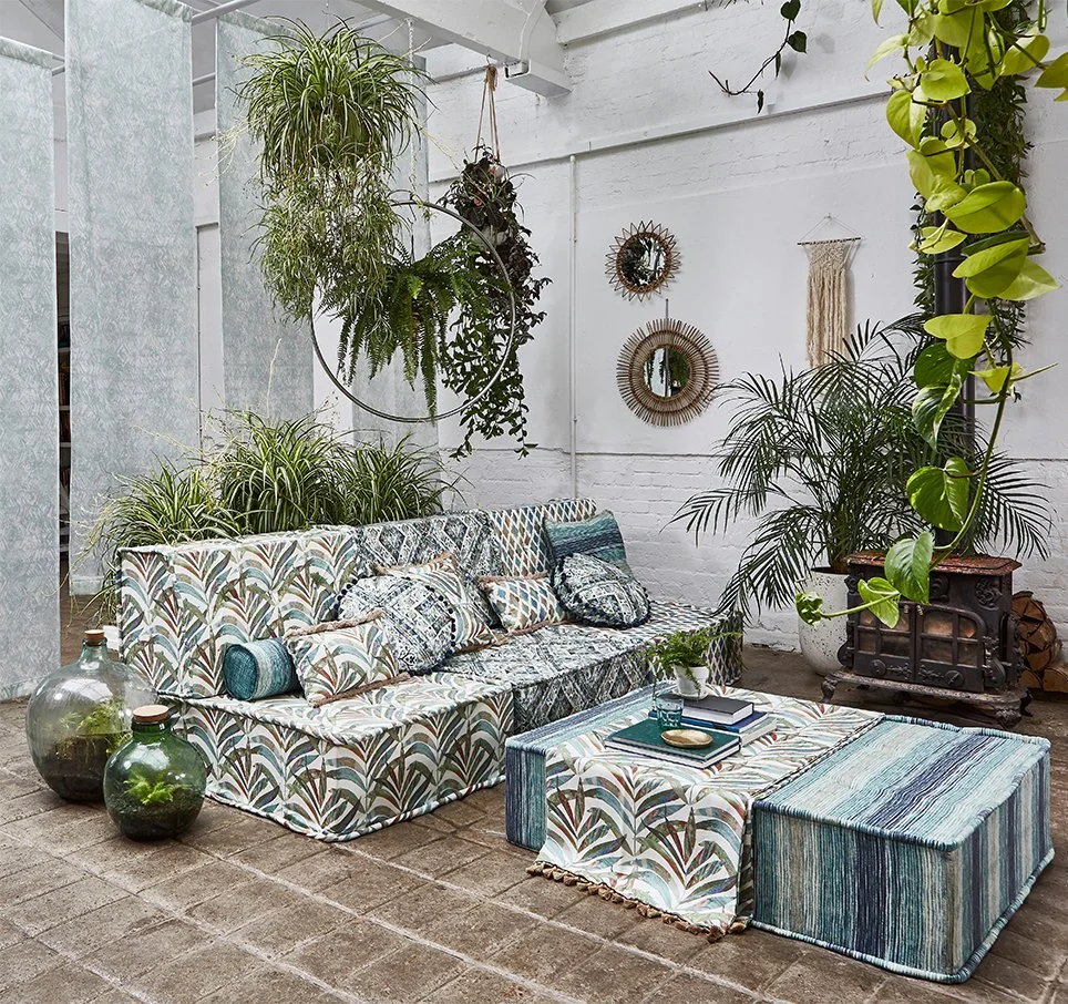 Bohemian-style indoor lounge with a patterned sofa, various pillows, large potted plants, hanging planters, mirrors, and a wooden stove against a white brick wall.