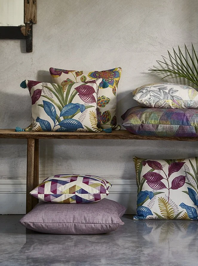Colorful decorative pillows with botanical leaf and plant patterns, arranged on a wooden bench and flooring next to a white wall and a green palm leaf.