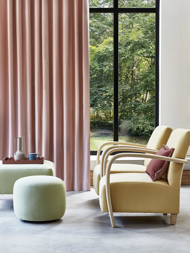 A cozy corner with two yellow armchairs, pink curtains, a green pouf, and a side table with decor, next to a large window showing green trees outside.