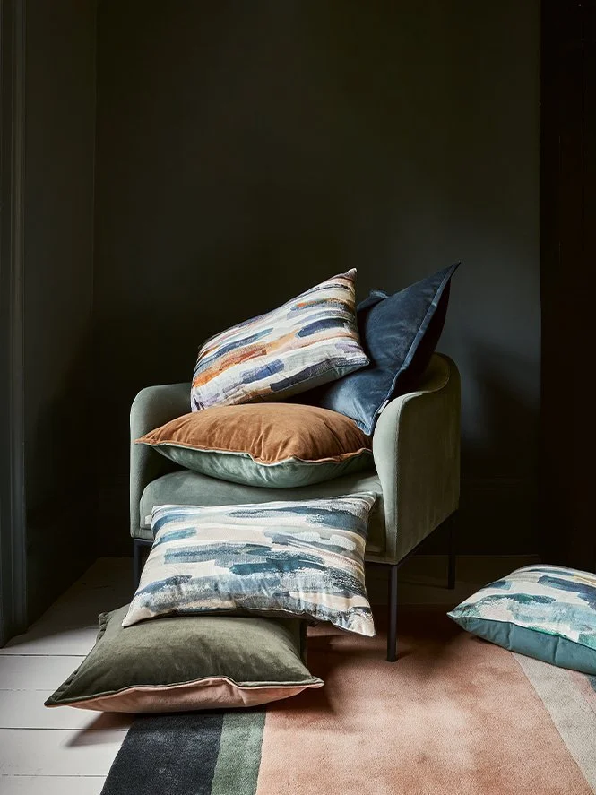A green armchair with multiple decorative pillows on and around it, set against a dark wall. The pillows are in various colors and patterns, with some on the chair and some on the floor and a rug.