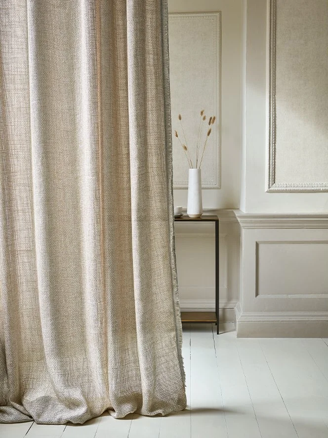 A beige curtain partially covering a white wall with decorative molding, a black metal and glass side table, and a tall white vase with dried plants inside.