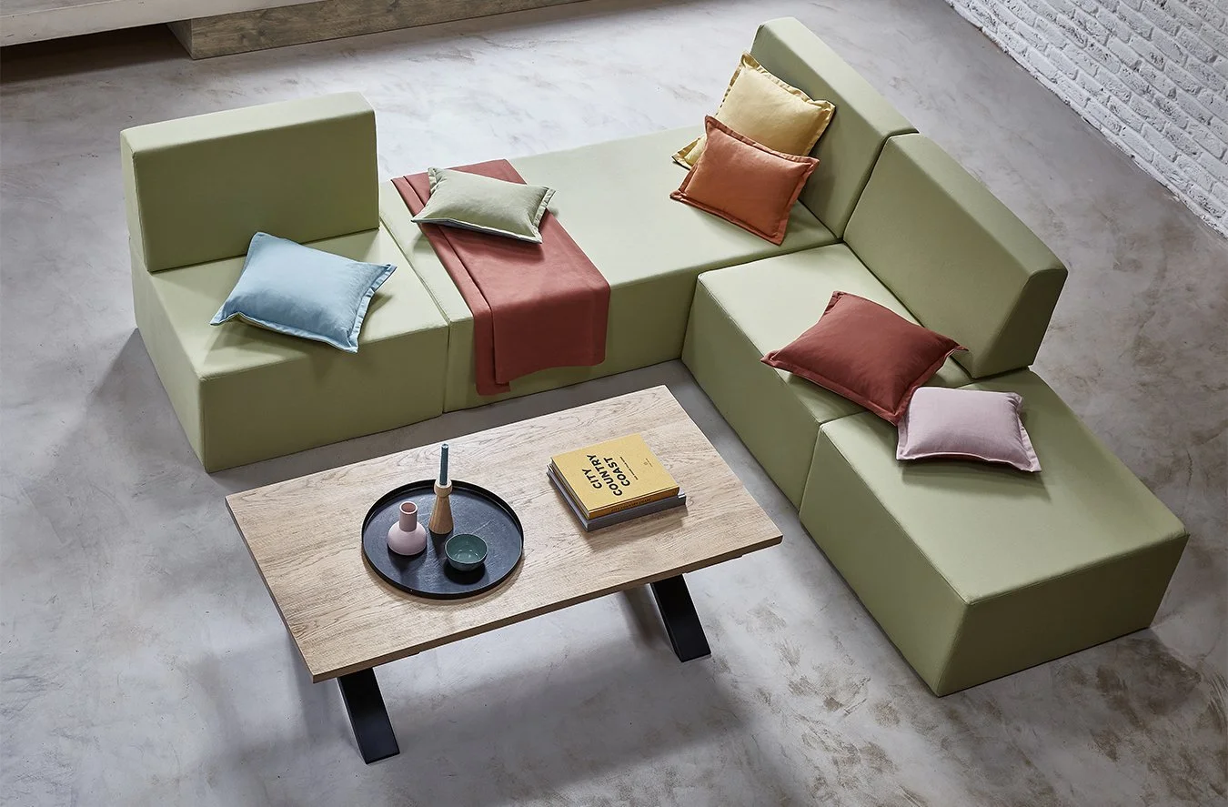 A modern green sectional sofa with multiple colorful throw pillows in a contemporary living room. A light wood coffee table with decorative vases and a yellow book is in front of the sofa. The room has a polished concrete floor and a white brick wall