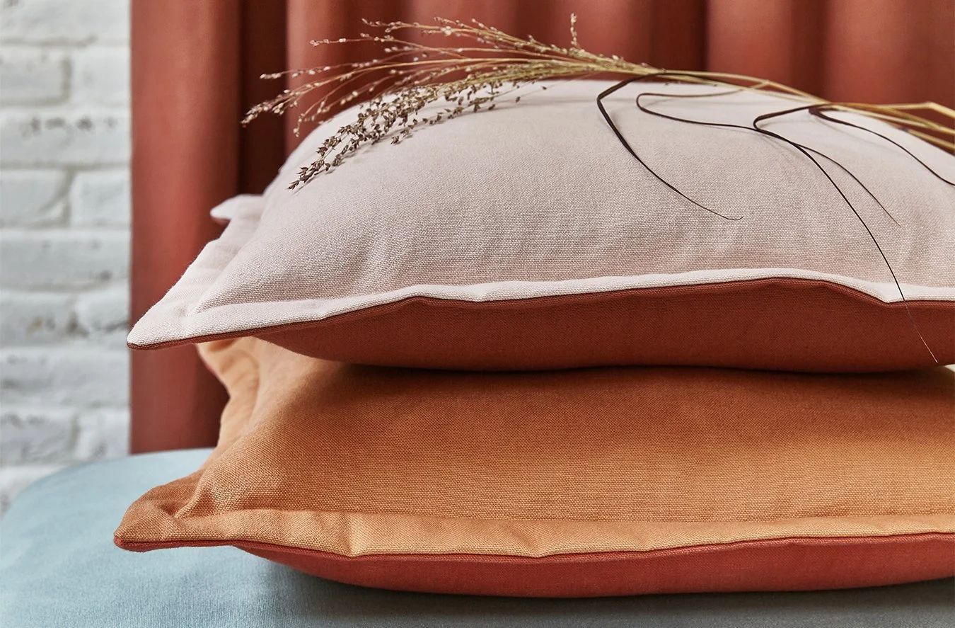 Stack of three pillows in earth tones on a blue surface with decorative dried plants on top. Background includes a white brick wall and rust-colored curtains.