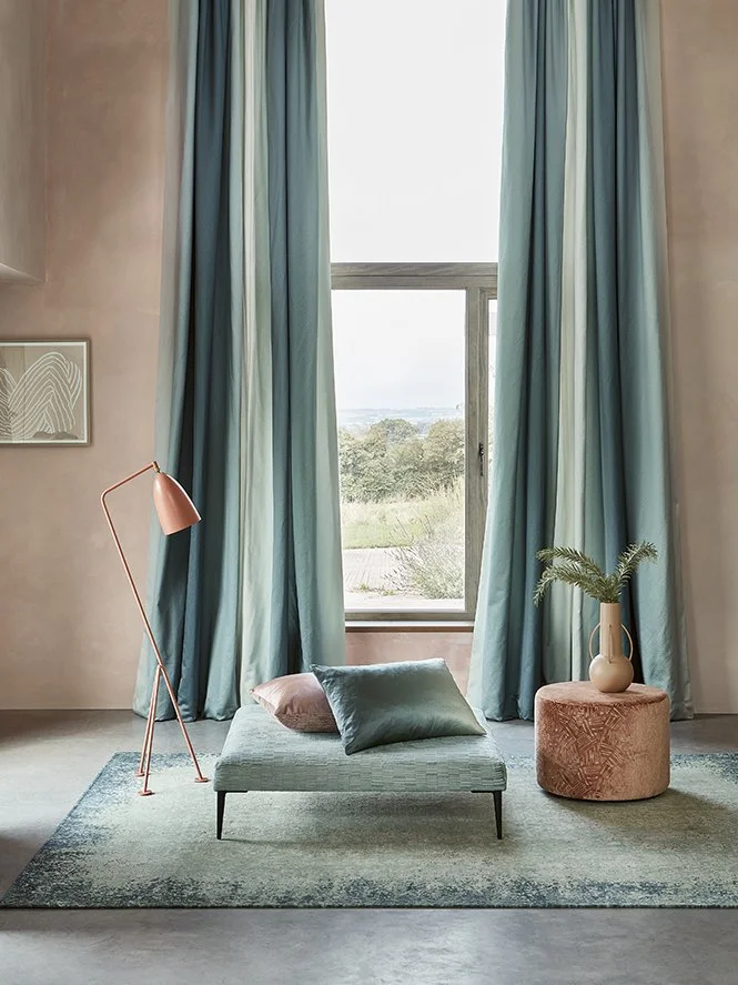 A cozy sitting area with a large window with blue curtains, a pink floor lamp, a low green ottoman with pillows, and a round beige ottoman with a potted plant.