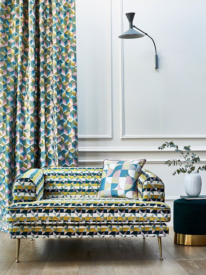 Interior living space with geometric patterned curtains, a matching patterned sofa with a throw pillow, a white wall with molding, a modern black and gray wall-mounted lamp, and a small black side table with a white vase and greenery.