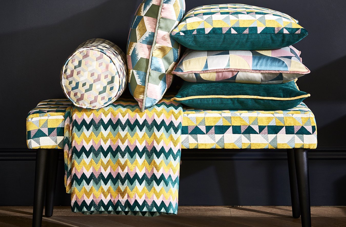 A bench with a colorful, geometric patterned cushion underneath a stack of matching patterned pillows, and a coordinating round pillow leaning against the stack, set against a black wall.
