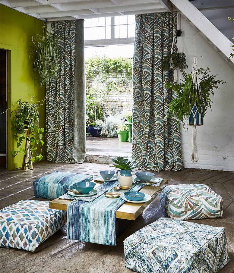 A cozy indoor dining area with a low wooden table, colorful patterned cushions and bowls, set near large open curtains leading to a lush outdoor garden with potted plants. Green accent wall and hanging plants add vibrancy.