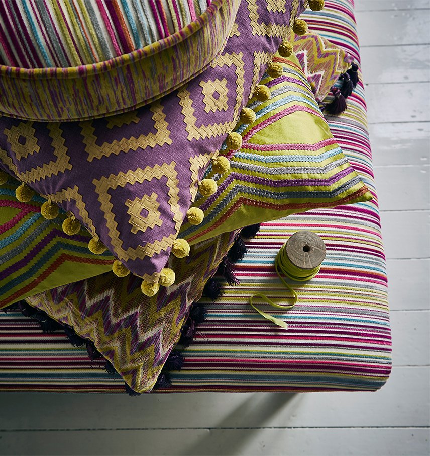 Colorful striped fabric and embroidered pillows on a white wooden bench, with a spool of green thread.