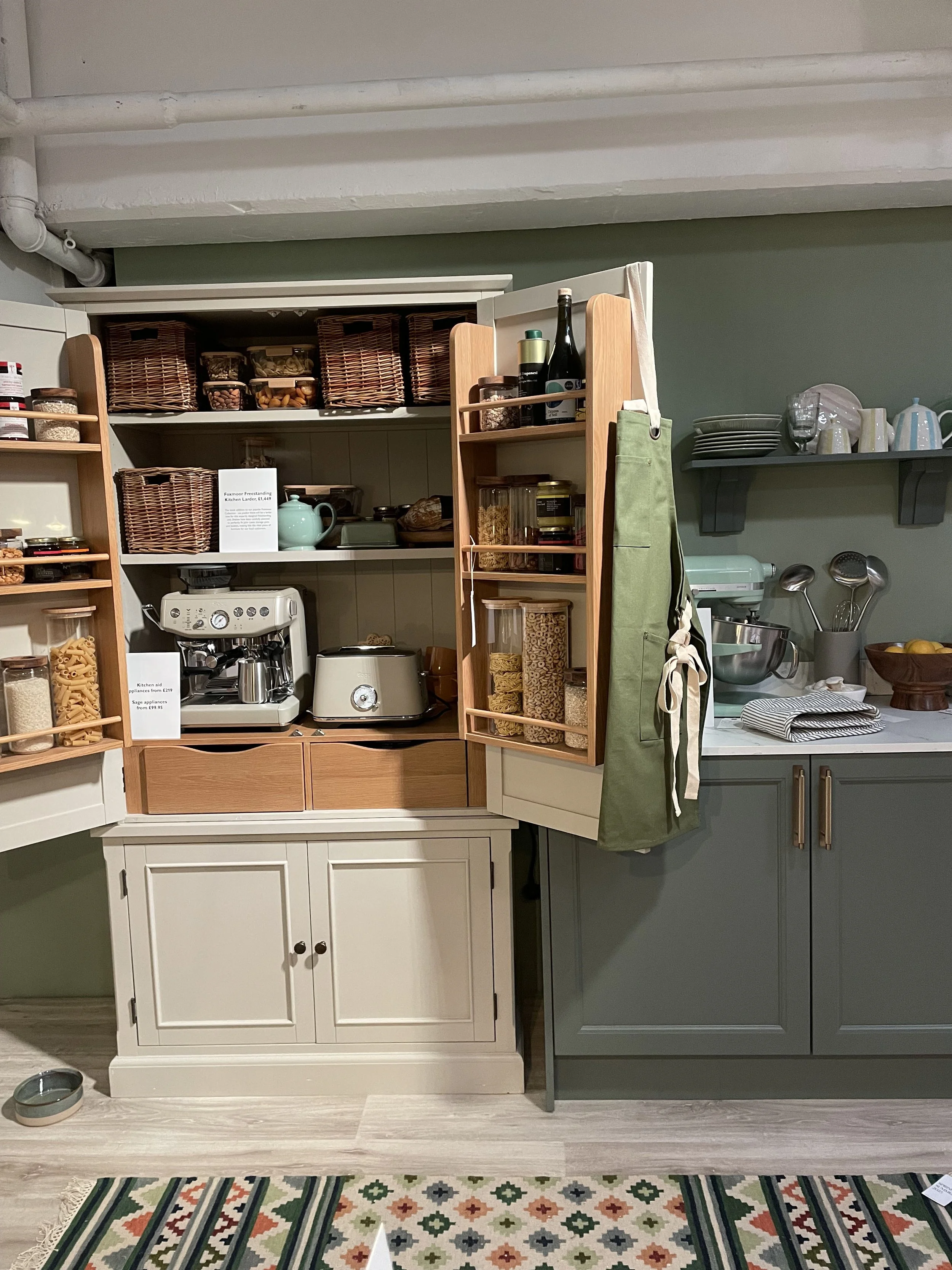 Kitchen cabinet filled with jars, containers, and appliances, with a green apron hanging on the door.