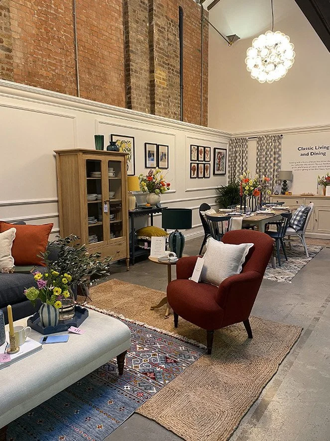 A cozy living room and dining area with decorated furniture, including a white upholstered sofa, a red armchair, a wooden cabinet, and a dining table with chairs. The room has decorative rugs, floral arrangements, wall art, and a modern light fixture