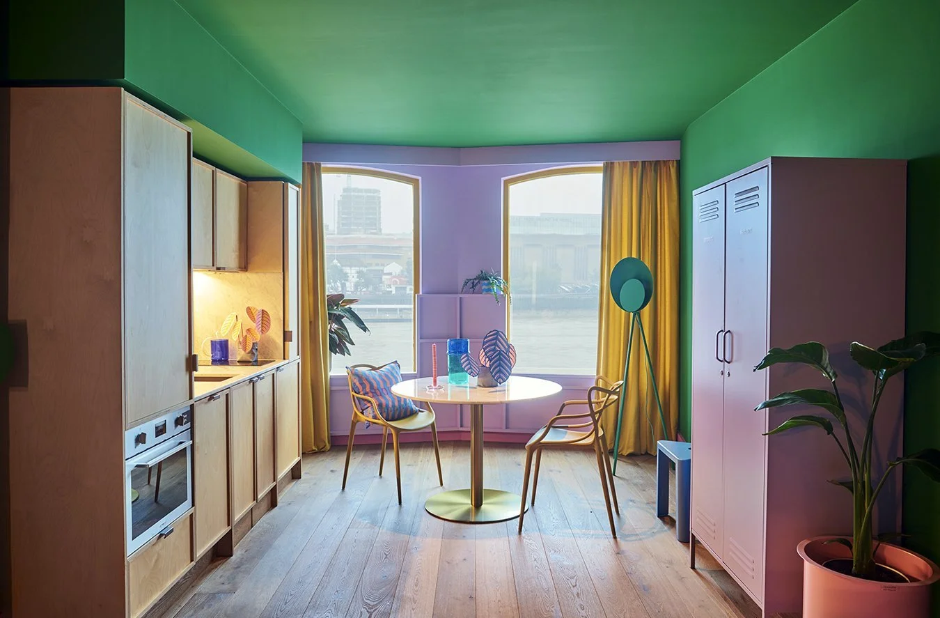 Bright, colorful kitchen with green ceiling and purple walls. Wooden cabinets, round table with yellow chairs, plants, large windows with yellow curtains, and modern decor elements.