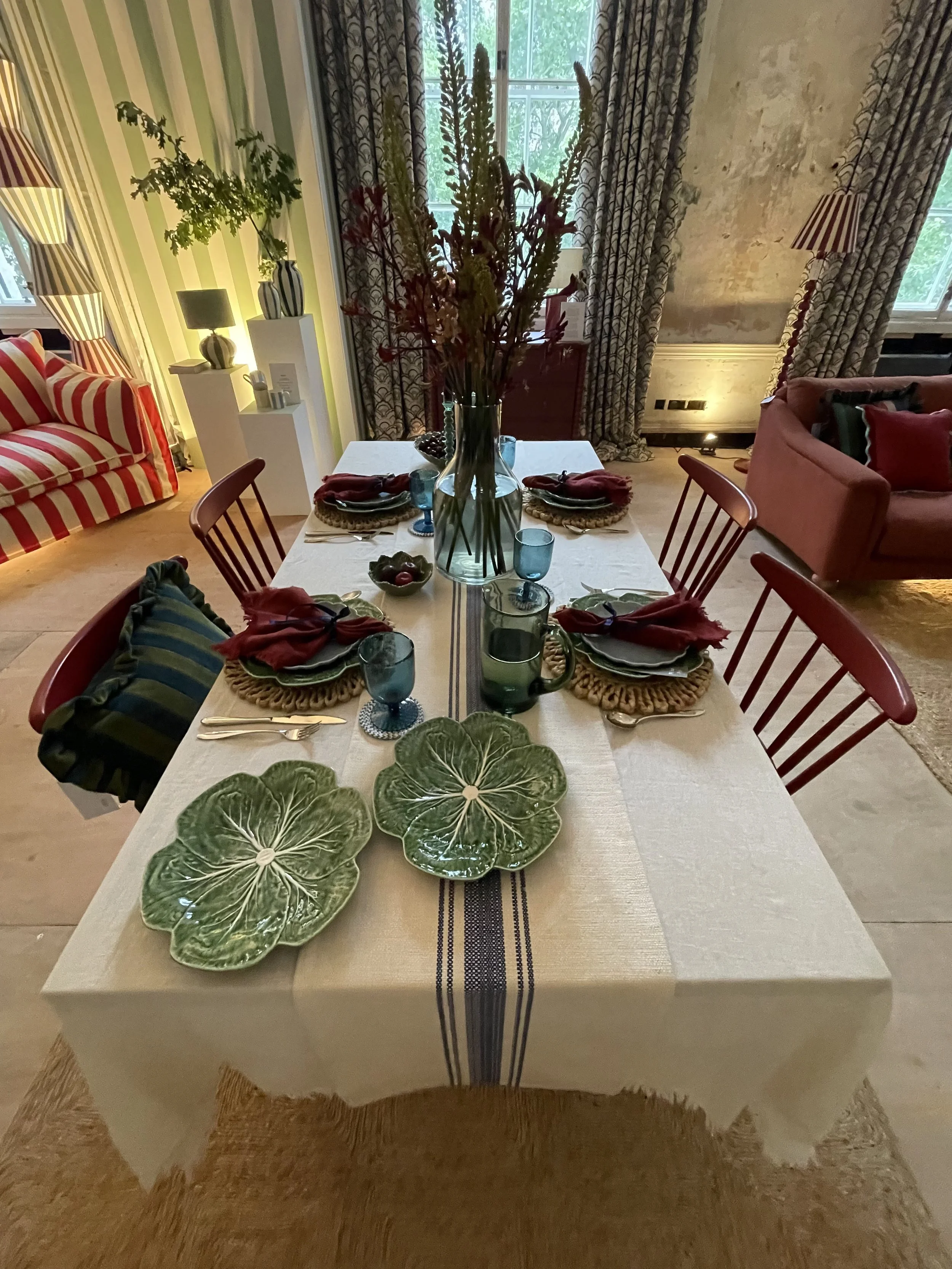 A dining table set for six with green leaf-shaped plates, red napkins, blue glasses, and a floral centerpiece with tall flowers in a glass vase. The room has patterned curtains, a striped sofa, a pink armchair, and decorative lamps.