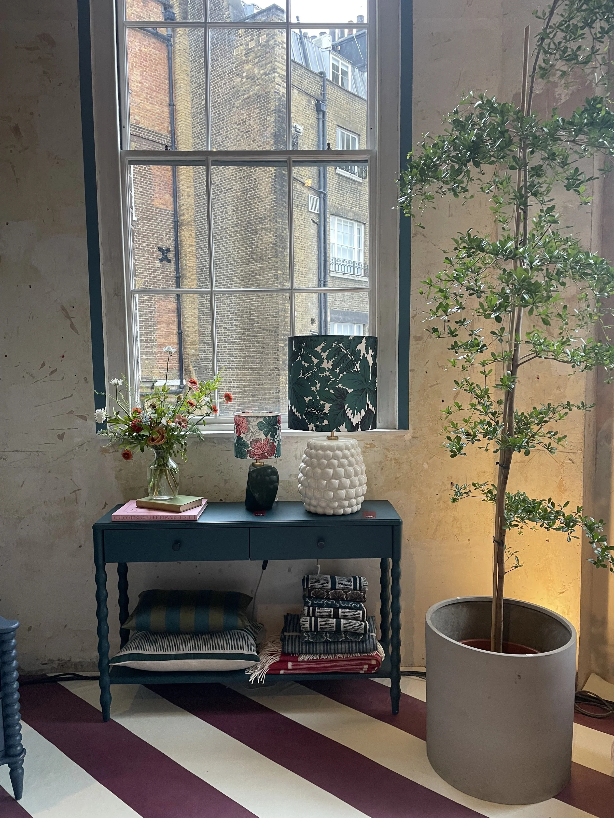 A blue side table with drawers, topped with floral lampshades, a flower vase, and books, against a window with view of brick buildings, with a large potted tree nearby.