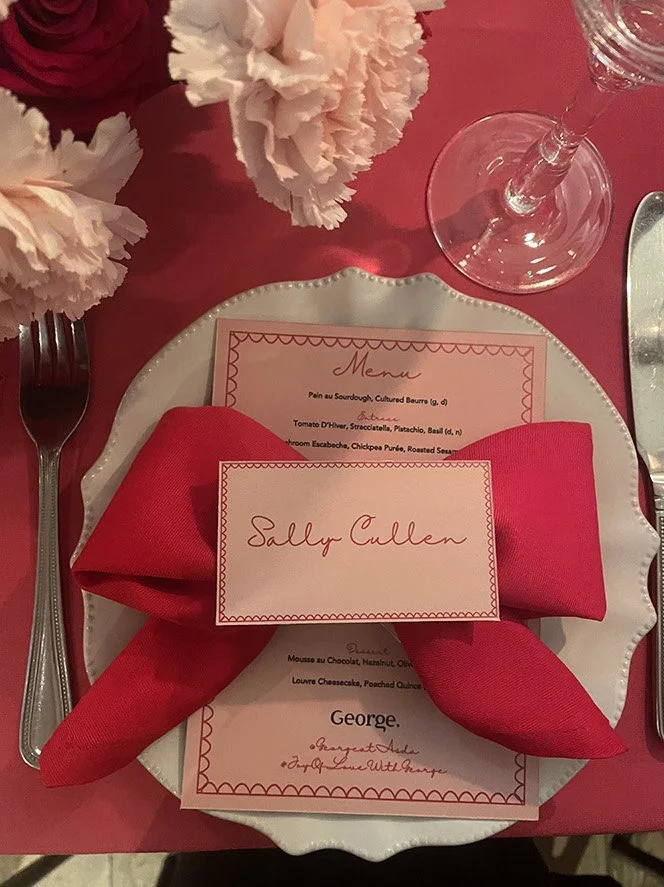 Place setting at a dining table with a white plate, a red napkin folded on the plate with a place card that reads "Sally Cullen," a menu underneath, a wine glass, and pink flowers in the background.