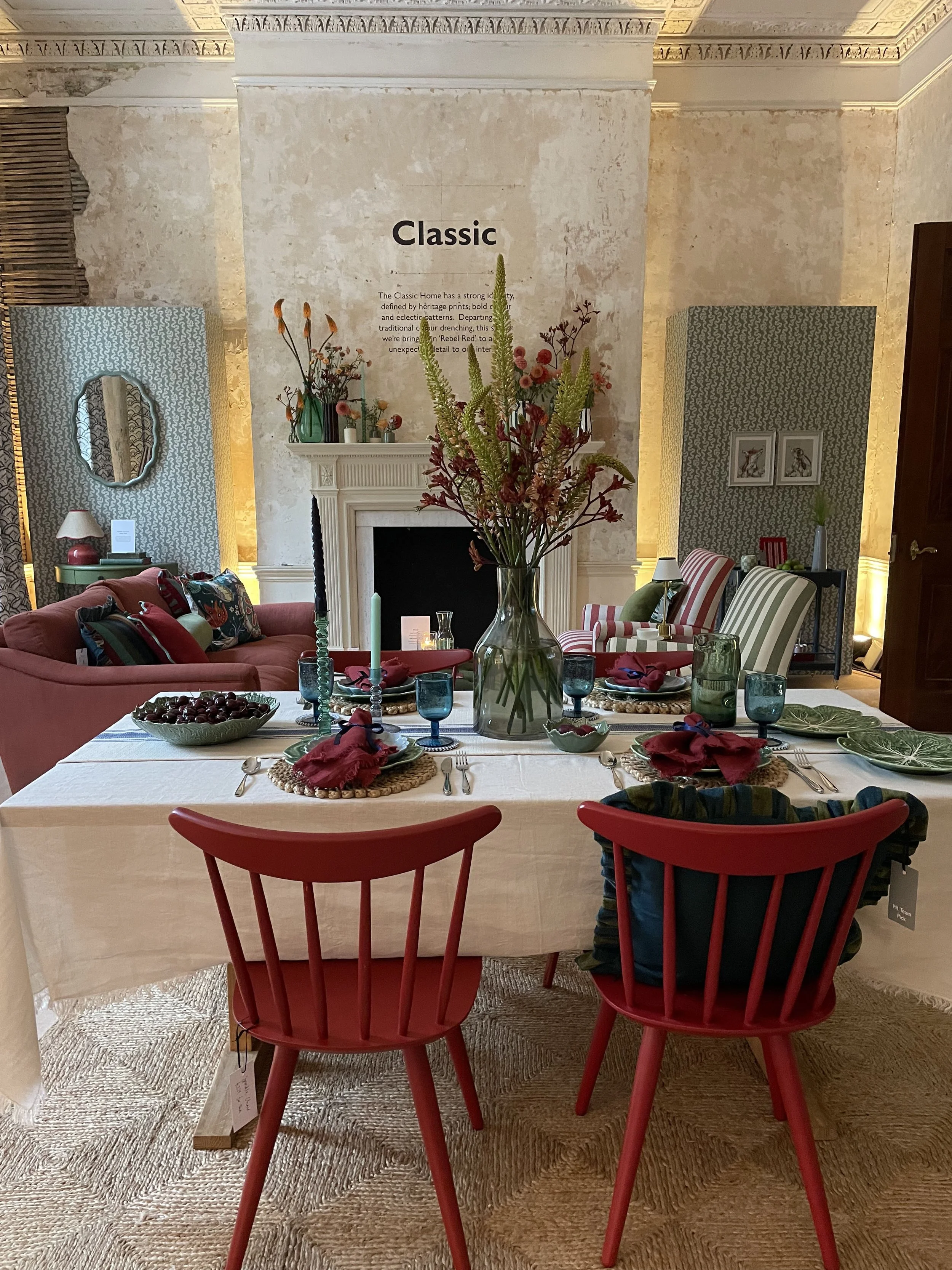 A dining room scene with a white dining table set with plates, glasses, silverware, and napkins, decorated with a large glass vase of pink and green flowers in the center. Four red chairs are around the table. In the background, a cozy living area wi
