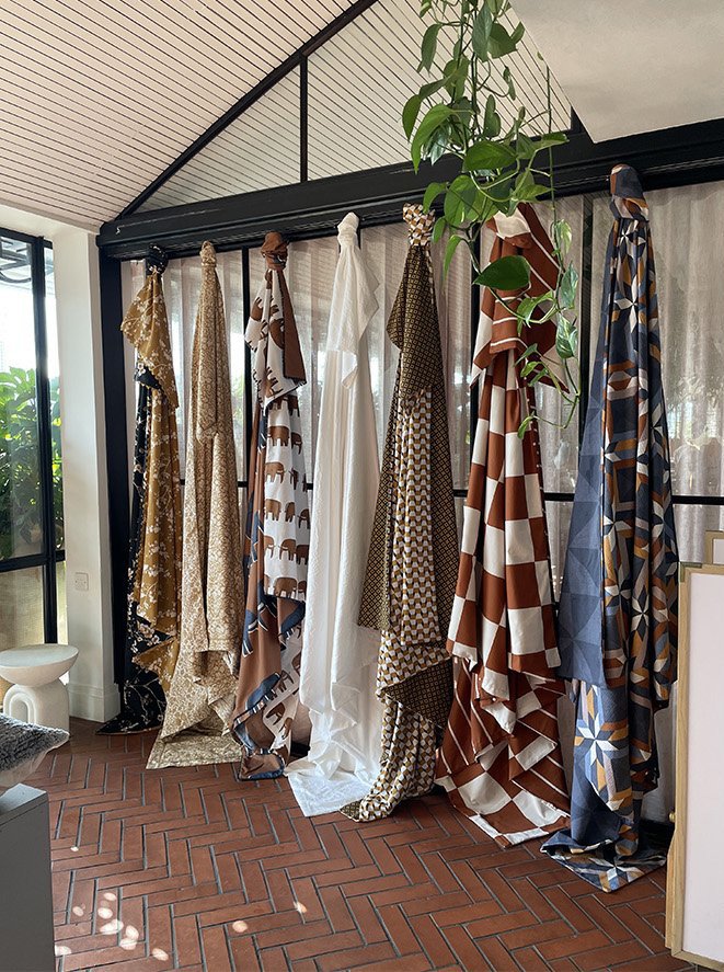 A display of various patterned and solid fabrics hanging on a black metal rack inside a room with brick tile flooring, a white wall, and a window with greenery outside.