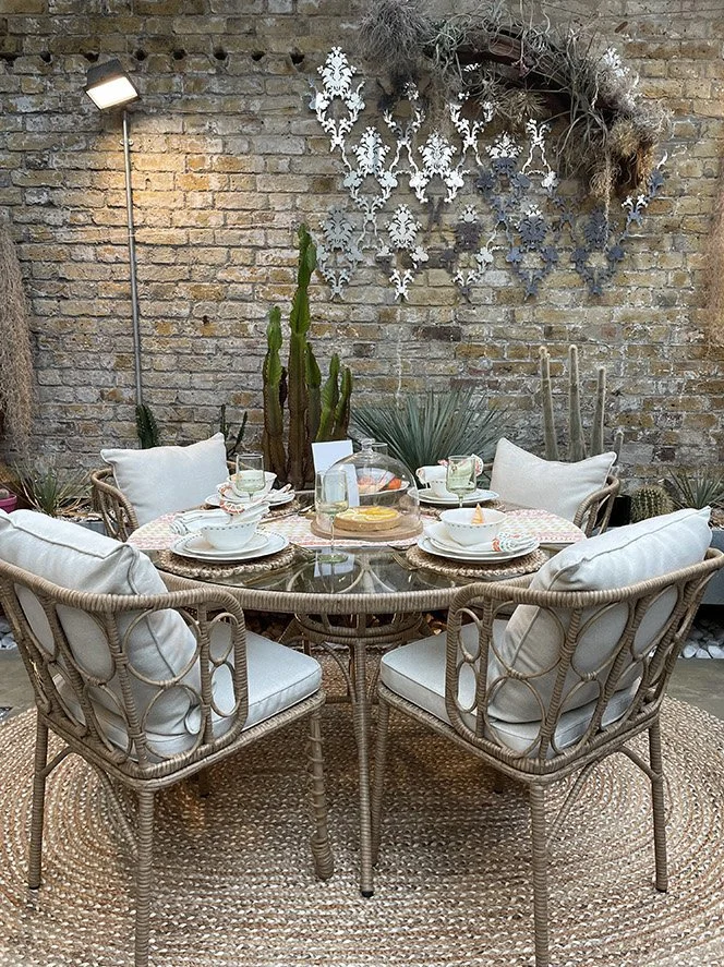 Round dining table set with six wicker chairs, white cushions, and tableware, situated on a braided rug against an exposed brick wall decorated with metal wall art and potted cacti.