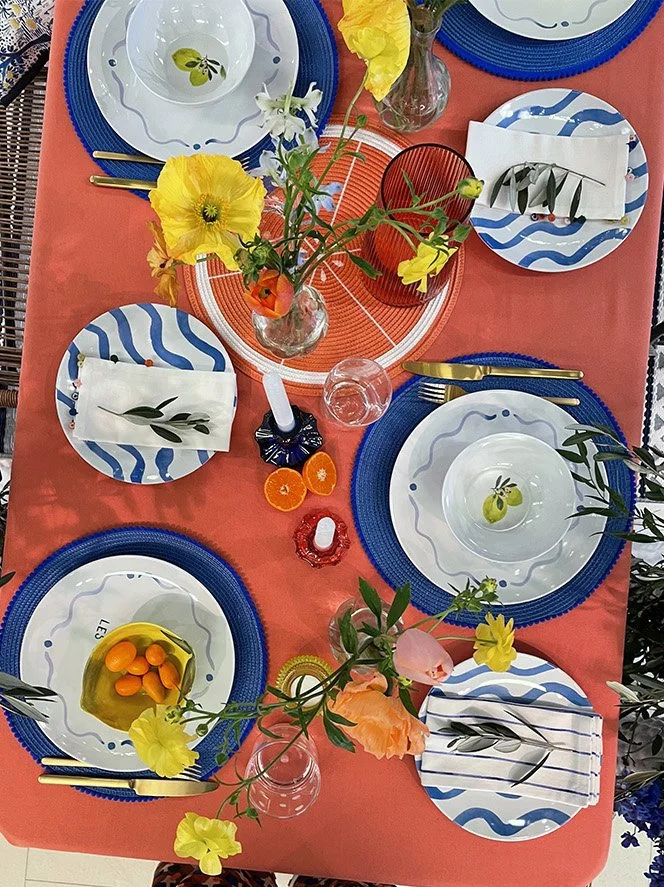 A colorful dining table set with patterned blue and white plates, gold utensils, floral napkins, and bright flowers in vases on a coral tablecloth.