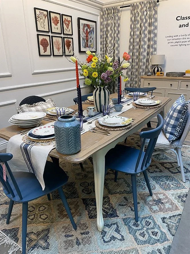 A dining room table set for a meal with a centerpiece of colorful flowers, blue and white tableware, and tall candle holders with red candles. Six chairs surround the table, three in dark blue and three in light blue and white. The room has a pattern