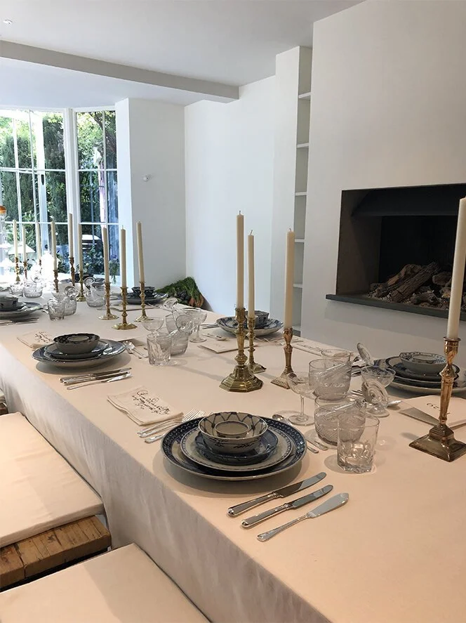 A long dining table set for a meal, with blue and white china, glassware, silverware, and tall beige candlesticks, in a bright room with large windows and a fireplace.