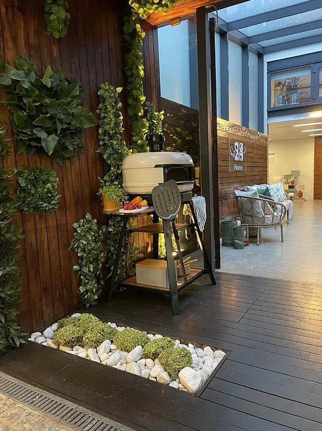 Indoor patio with wooden flooring, green plants on a wooden wall, a small grill on a stand, a table with a bowl of fruit, and a seating area with cushions.