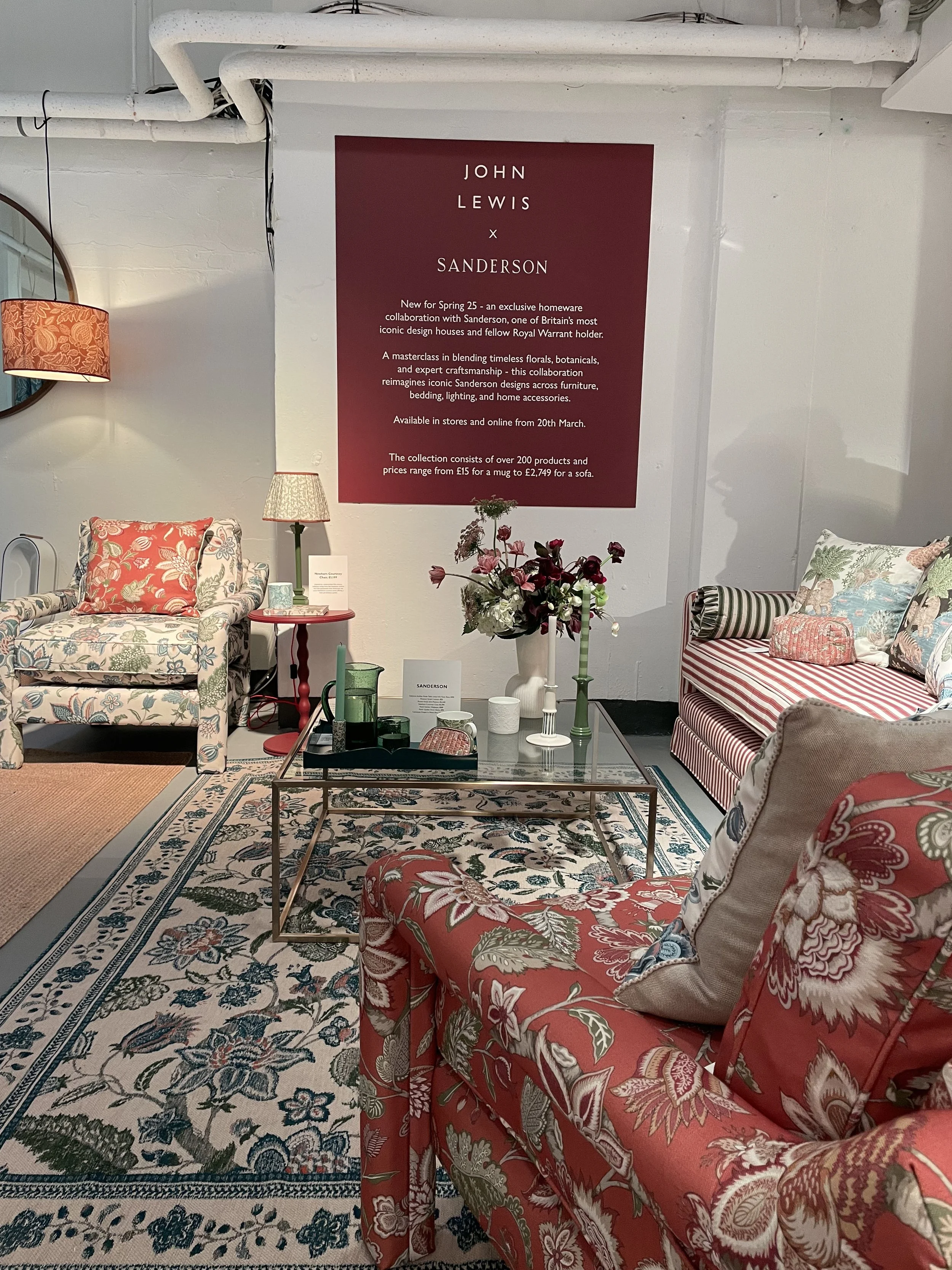Living room setup with floral-patterned armchairs, sofas, a glass coffee table, and decorative vases, with a large promotional poster for a collaboration between John Lewis and Sanderson on the wall.