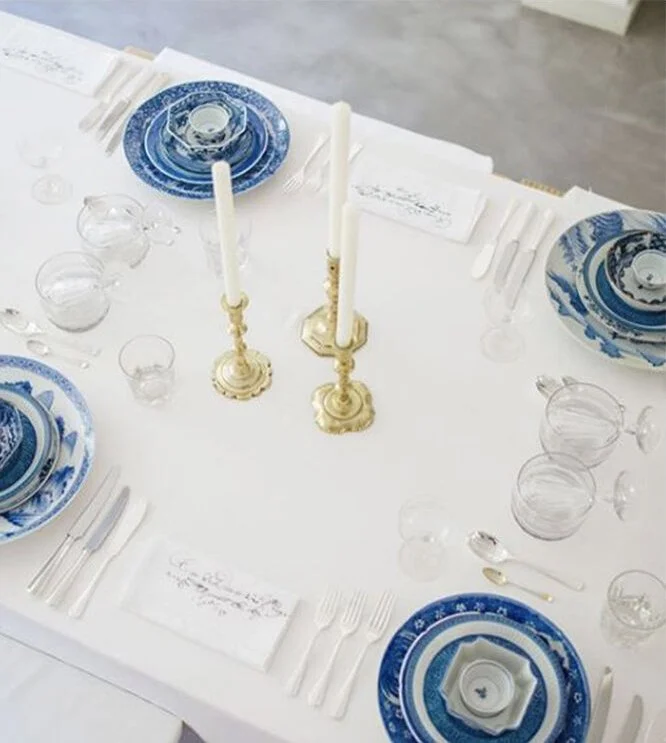Table setting with blue and white patterned plates, clear glassware, white napkins, and two tall white candles in gold candleholders on a white tablecloth.