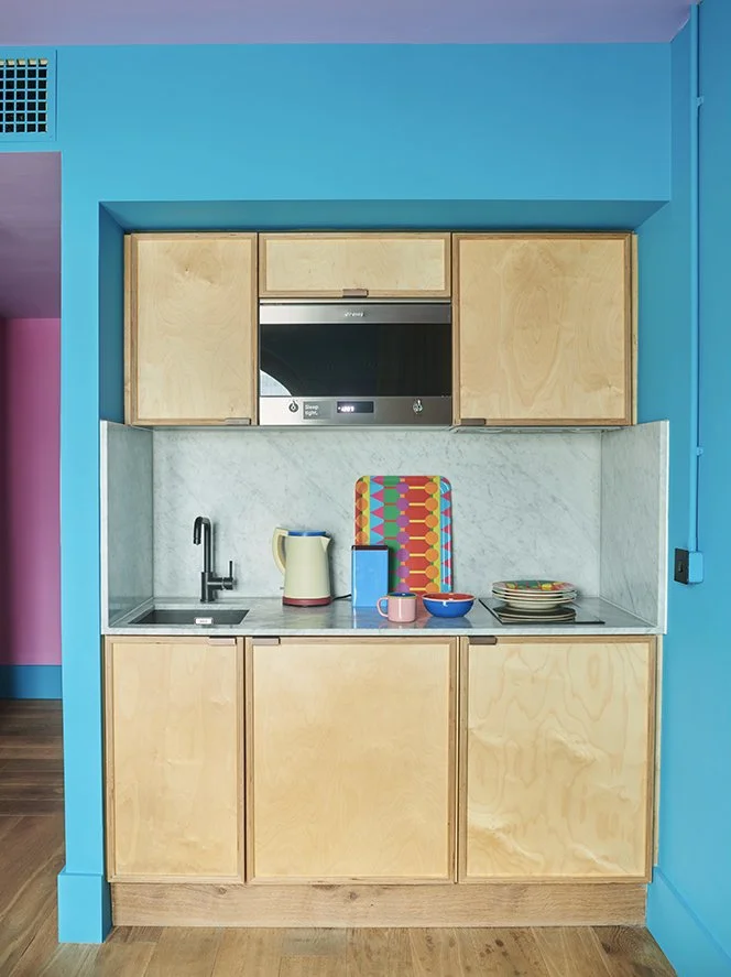 Small kitchen with light wood cabinets, a marble backsplash, black faucet, electric kettle, colorful tray, mug, bowl, and plates.