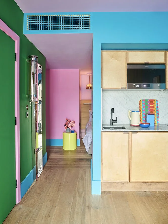 Colorful kitchen with green wall and pink door frame, pastel blue wall, yellow cabinets, marble countertop, microwave, kettle, bowl, and decorative tray, with a bedroom visible in the background.