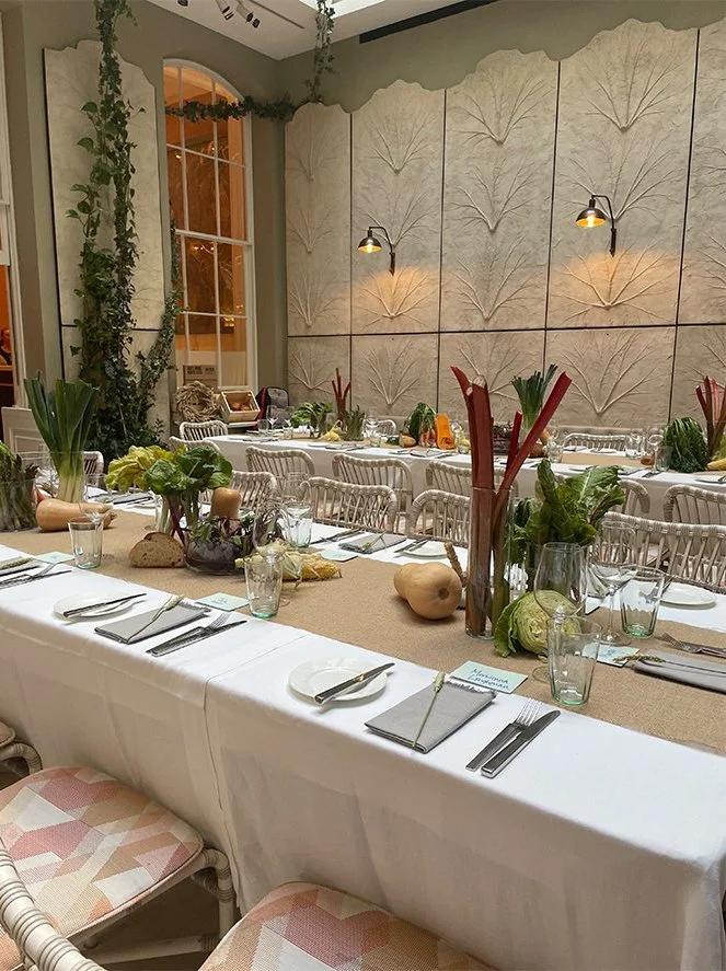 A long dining table set for a gathering with white plates, cutlery, and glasses, decorated with vegetables and greenery, in a room with high ceilings, large windows, and textured wall panels.