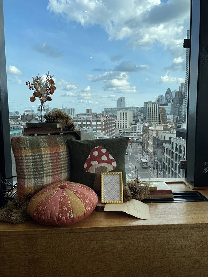 View of a city skyline through a window, with decorative items on the windowsill including cushions, a framed picture, a book, a dried plant arrangement, and a small vase with dried flowers.